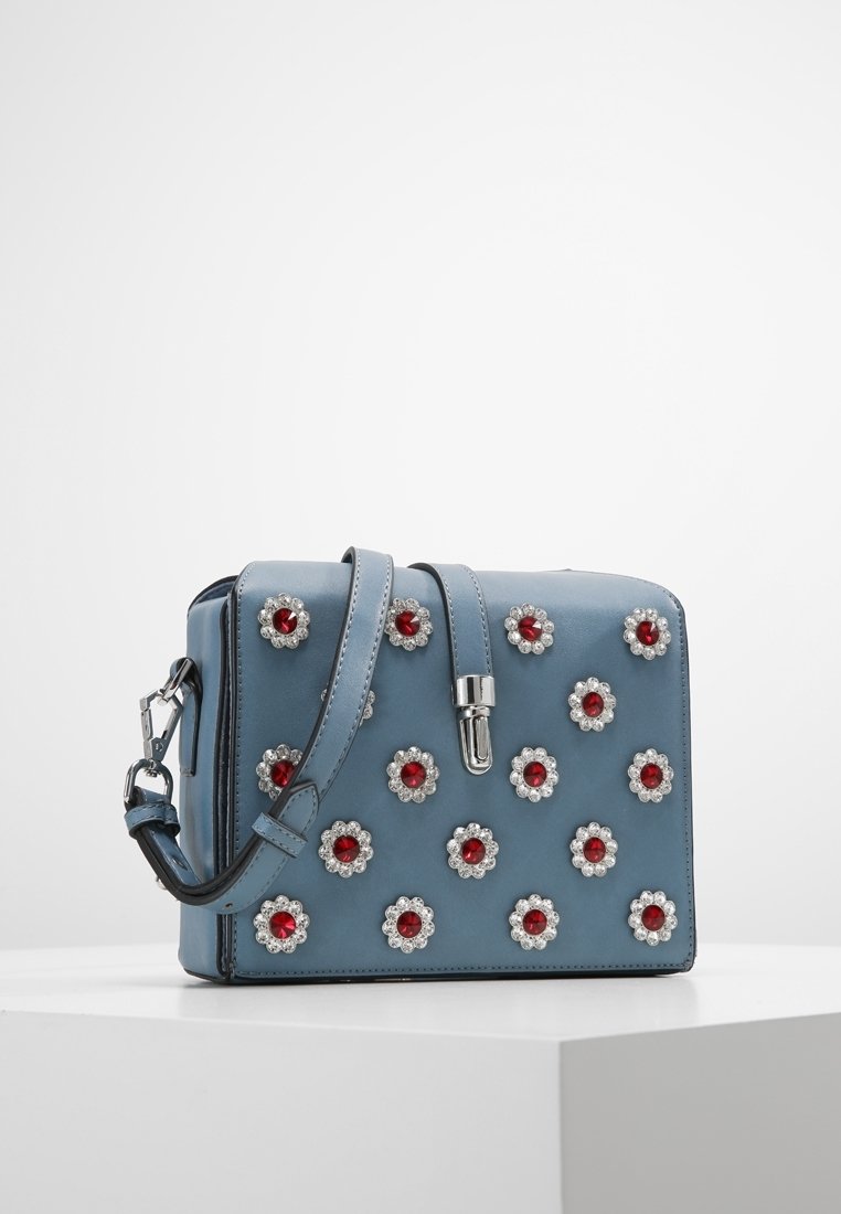 Sac à main en cuir bleu avec des embellissements floraux aux centres rouges et des accents argentés, bandoulière réglable et forme rectangulaire structurée.