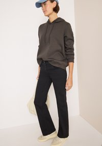 Felpa grigia scura con coulisse, abbinata a jeans neri a zampa d'elefante. Sneakers beige e un cappellino chiaro completano l'outfit casual. Si notano texture morbide.