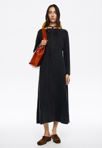 Vestido negro de manga larga con cuello alto, costuras verticales y un acento marrón. Zuecos de ante marrón; bolso naranja con correa.