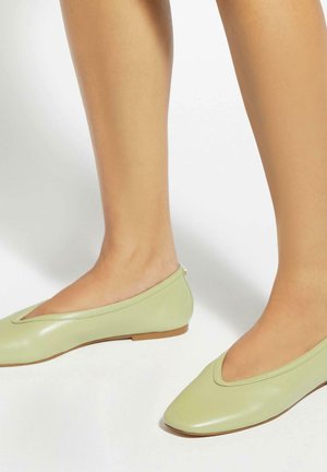 Benen met lichtgroene balletschoenen met een lage hak, weergegeven tegen een effen witte achtergrond.