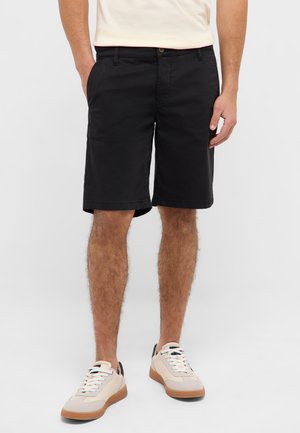 Mann trägt schwarze knielange Shorts, beige Turnschuhe mit weißen Schnürsenkeln und ein helles Hemd, steht mit den Händen in den Taschen.