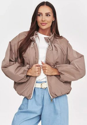 Kobieta z długimi ciemnymi włosami nosi beżową oversize kurtkę na suwak, biały top, niebieskie spodnie z plecionym paskiem, spogląda na bok.