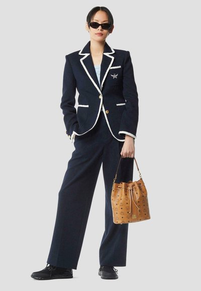 Vrouw draagt een donkerblauw pak met witte bies, zwarte sneakers, zonnebril, en houdt een bruine, met patroon beschilderde emmervormige tas vast tegen een effen achtergrond.