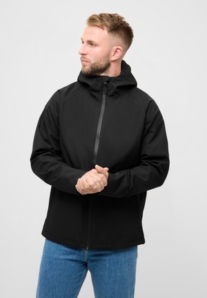 Mand med kort blondt hår iført en sort hættetrøje og blå jeans, der kigger mod venstre med hænderne foldet foran sig.