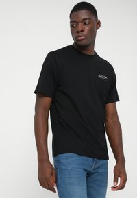 Μαύρη βαμβακερή μπλούζα με κοντά μανίκια και στρογγυλή λαιμόκοψη, με μικρό λευκό λογότυπο στην επάνω αριστερή πλευρά, φορεμένη με μπλε τζιν.