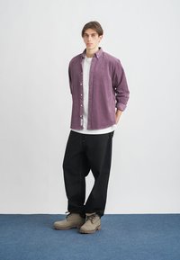 Fioletowa koszula z kołnierzykiem z korduroju na białym t-shircie, połączona z czarnymi, luźnymi spodniami i beżowymi botkami za kostkę na niebieskiej podłodze.