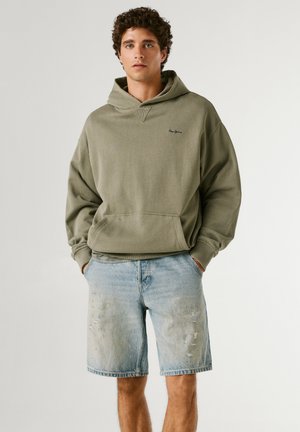 Ung mand iført en olivengrøn hættetrøje og lyseblå slidte denimshorts med hænderne i lommerne mod en ensfarvet baggrund.