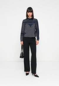 Langärmlige, marineblaue Bluse mit weißen geometrischen Linien, kombiniert mit schwarzen Hosen. Schwarze spitze Pumps und eine kleine schwarze Handtasche.