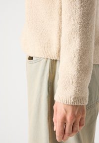 Main d'une personne à côté de la manche d'un pull beige texturé et d'un pantalon en denim bleu clair visible de la taille jusqu'aux doigts.