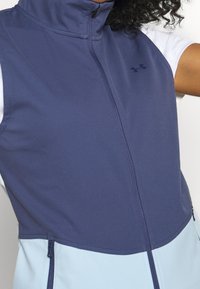 Navy blue sleeveless vest with a high collar, zip closure, and light blue bottom panel; features Under Armour logo and functional side pockets.