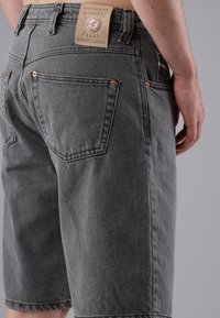 Close-up of a person wearing gray denim shorts with a tan leather label on the waistband reading "Picaldi Jeans Authentic Design."