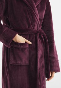 Accappatoio morbido di un profondo colore viola con collo a scialle, cintura da annodare e tasca frontale. Texture soffice con polsini a coste.