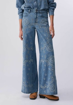 Niebieskie dżinsy z szerokimi nogawkami i wysokim stanem, ozdobione białym haftem w stylu paisley, noszone z dopasowaną haftowaną jeansową koszulą i brązowymi botkami na obcasie.