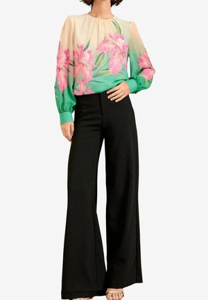 Femme portant un chemisier à manches longues à motif floral avec des fleurs roses et des feuilles vertes, associé à un pantalon noir taille haute à jambes larges.