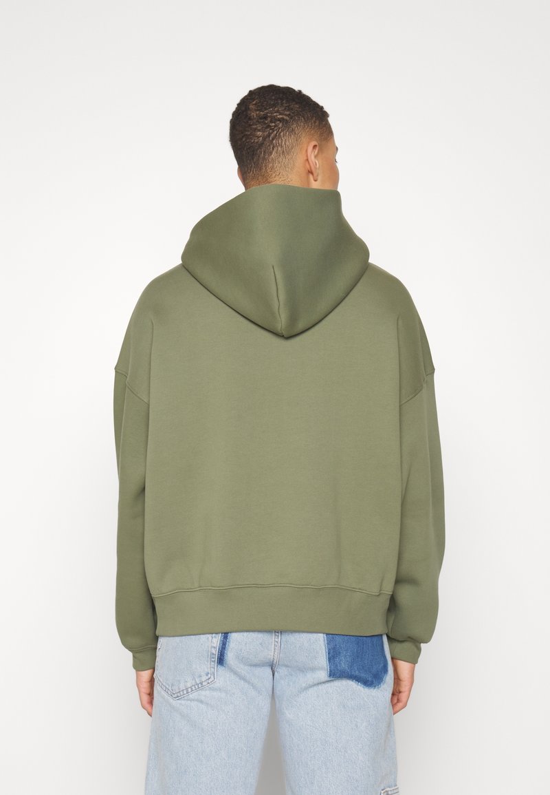 Homme de dos portant un sweat à capuche oversize vert olive et un jean bleu clair avec un patch bleu foncé sur la poche arrière.