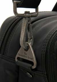 Nahaufnahme einer schwarzen Ledertasche mit einem Metallclip und einem Anhänger. Der Clip ist silberfarben und hat eine strukturierte Oberfläche, er ist an einem glatten Griff befestigt.