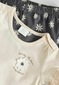 Camiseta de bebé en color crema claro con bordado floral y cierre a presión, combinada con pantalones cortos gris oscuro con patrones de margaritas blancas.