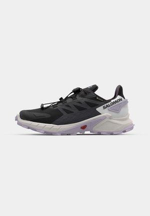 Zapatilla de trail running negra con un upper texturizado, suela flexible y un distintivo acento púrpura en el talón. Presenta un sistema de lazado rápido.