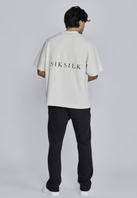 Gaiši bēšs īsām piedurknēm T-krekls ar lielu melnu "SIKSILK" logo aizmugurē; apvienots ar melnām biksēm un baltām kedām.