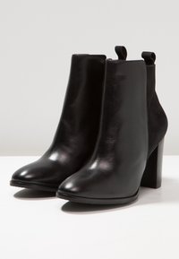 Botas negras de tobillo con un acabado de cuero liso, punta afilada y tacón cuadrado. Presentan un detalle de costura y tiradores en la parte trasera.