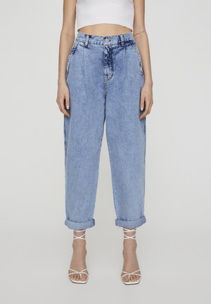 Kobieta w wysokich, jasnoniebieskich jeansach z podwiniętymi nogawkami, białych sandałach na obcasie z paseczkami oraz krótkim, białym topie bez rękawów, stojąca na jednolitym tle.