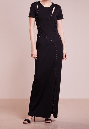 Femme portant une robe noire longue au sol à manches courtes, avec des ornements cloutés courbés et des découpes latérales, associée à des talons ouverts noirs.