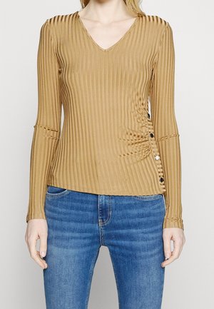 Kvinde iført en ribbet, tanfarvet langærmet top med V-hals og knapdetalje på højre side, kombineret med blå jeans.