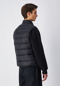 Schwarze Steppweste, gestepptes Design mit horizontalen Abschnitten, hoher Kragen, leichtes Material. Trägt man über einem schwarzen Langarmshirt.