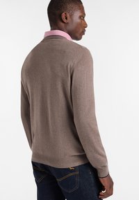 Maglione lavorato a maglia marrone con orlo e polsini a coste. Il colletto presenta una fodera contrastante rosa. Indossato sopra jeans blu scuro.