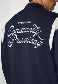 Marinejakke med hvite detaljer, med brodert tekst "STUDENTER Som Hele Tiden Øver" på baksiden i kursiv skrift. Glatt tekstur.