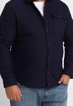 Mężczyzna ubrany w ciemnogranatową pikowaną koszulę zapinaną na guziki, nosi ją na białej koszulce, do tego jasnoniebieskie dżinsy, jedna ręka w kieszeni.