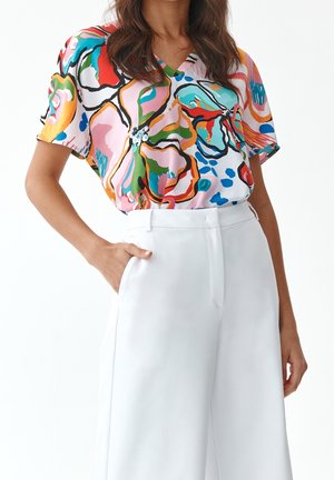 Kleurige blouse met korte mouwen op een roze achtergrond, met een bloemenpatroon in rood, groen en blauw. Gepaard met op maat gemaakte witte broeken.