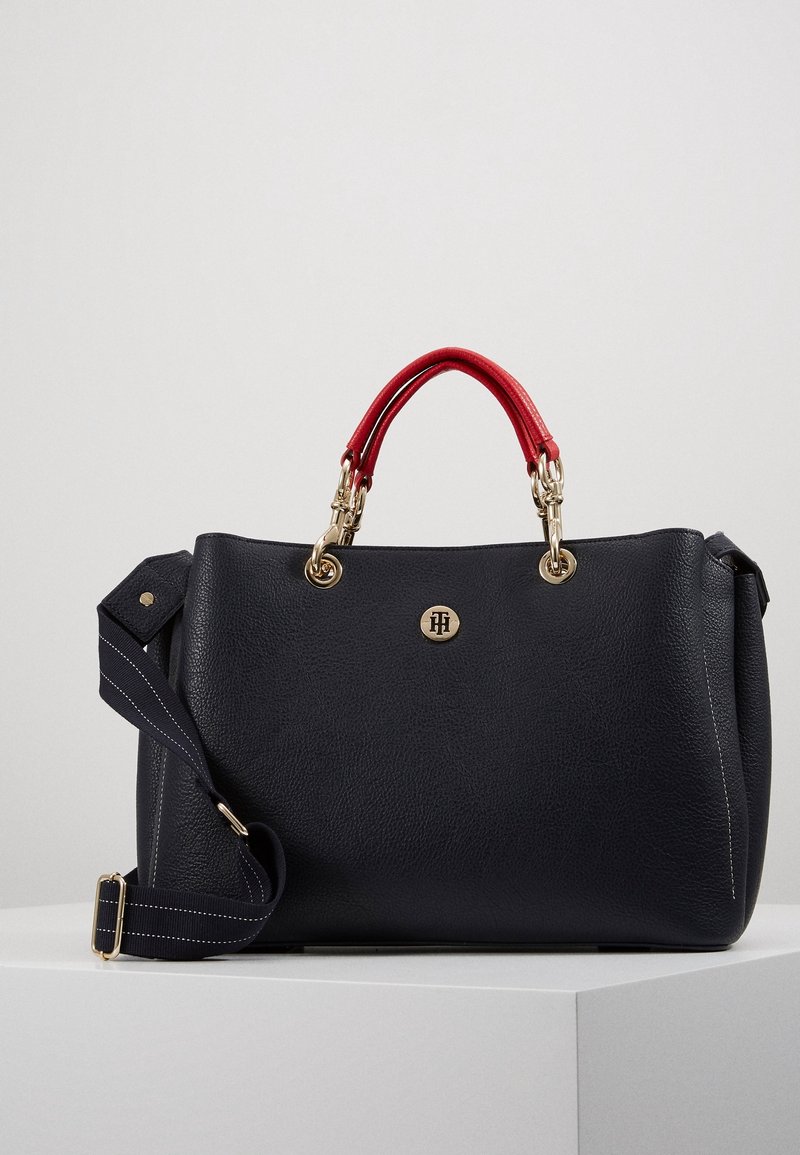 Czarna skórzana torebka z kontrastującymi czerwonymi uchwytami, złotymi detalami i odpinanym niebieskim paskiem w paski. Logo na froncie. Gładka faktura.