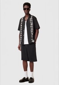 Camisa preta de manga curta com texto branco, combinada com calções pretos. Modelo usa óculos de sol, t-shirt branca, meias brancas e sapatos pretos.