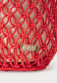 Virkat väska i ljusröd med nätmönster, med en tygbotten. Inkluderar en etikett med "HVISK" i vitt på en tan bakgrund.