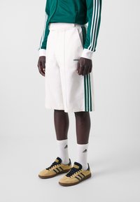 Vita shorts med gröna ränder, kombinerade med gula sneakers med svarta detaljer. Modellen bär en grön zip-up jacka och vita strumpor.