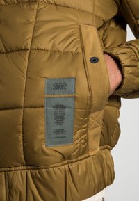 Olivgrön puffjacka med kviltade sektioner, sidoknappar och dubbla tygetiketter med skötsel- och märkesinformation.