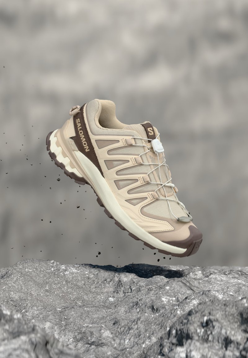 Chaussure de trail running Salomon beige et marron en l'air au-dessus d'une surface rocheuse avec de petites particules de terre tombant autour.
