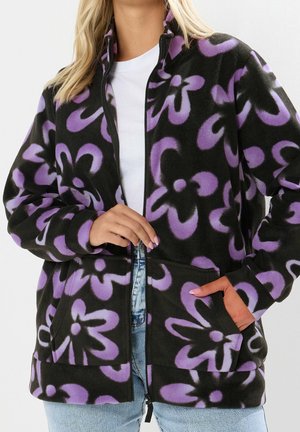 Kobieta ubrana w czarną kurtkę polarową z dużymi fioletowymi kwiatowymi wzorami, zapinaną do połowy, ręce z przodu i w kieszeni, w połączeniu z jasnoniebieskimi jeansami.