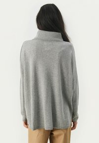 Donna con lunghi capelli scuri che indossa un ampio maglione grigio a collo alto e pantaloni beige, vista di spalle su uno sfondo bianco.