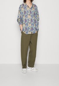 Camisa de manga larga colorida con estampado floral en tonos de púrpura y beige, combinada con pantalones sueltos color verde olivo y zapatillas blancas.