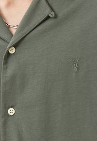 Olivengrønn t-skjorte med korte ermer, laget av lettvevde materialer, med en notched krage, to knapper, og en subtil brodert geite-logo.