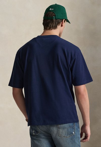 Μπλε ναυτικό βαμβακερό μπλουζάκι με κοντά μανίκια, στρογγυλή λαιμόκοψη και άνετη εφαρμογή. Φορεμένο με ανοιχτό μπλε τζιν με φθορές και πράσινο καπέλο.