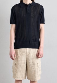 Námořnická pletená polo košile s krátkými rukávy, s texturovaným vzorem a uzávěrem na tři knoflíky. V kombinaci s béžovými cargo šortkami.