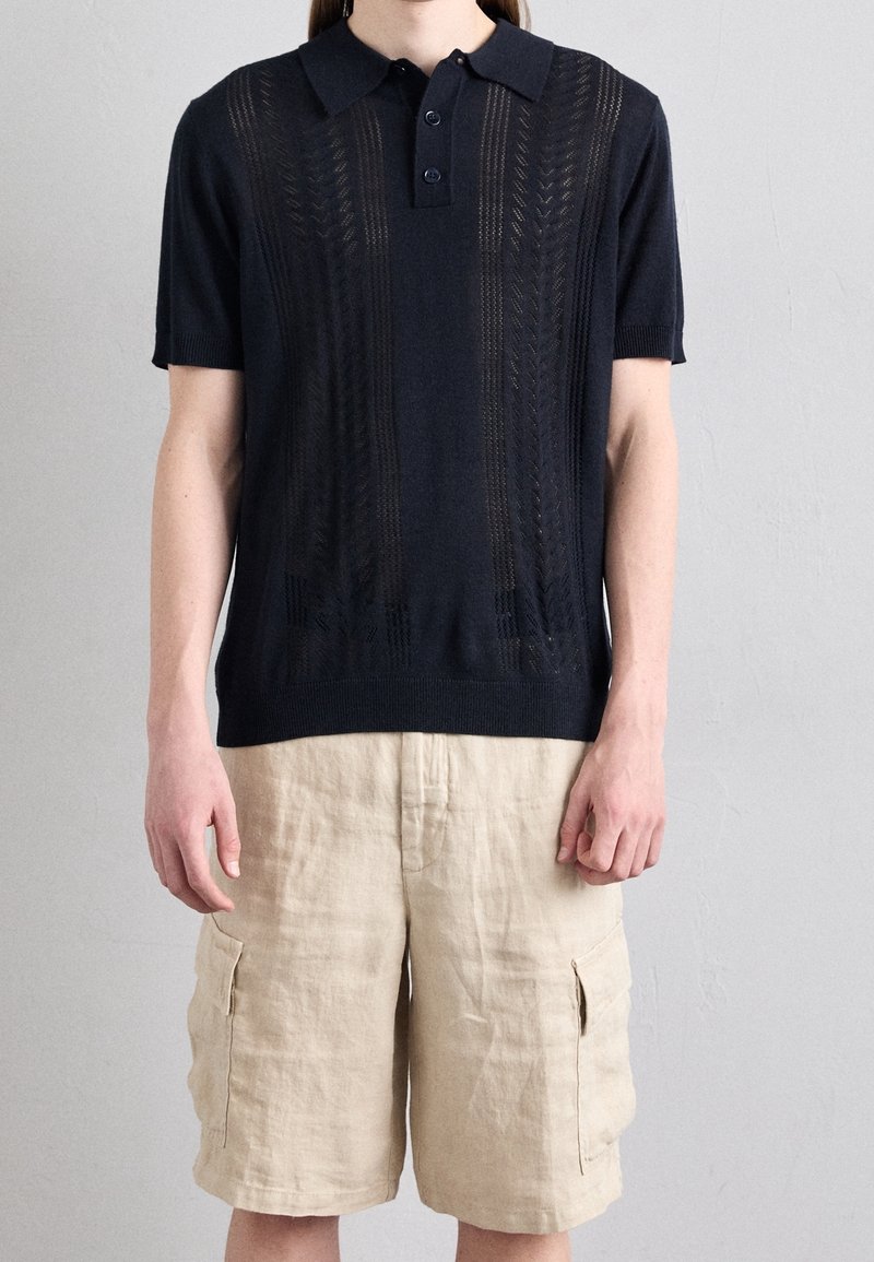 Námořnická pletená polo košile s krátkými rukávy, s texturovaným vzorem a uzávěrem na tři knoflíky. V kombinaci s béžovými cargo šortkami.
