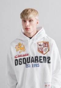 Jonge man met blond haar die een witte hoodie draagt met een gele leeuw, een wapen met cijfers en de tekst "DSQUARED2 EST 1995".