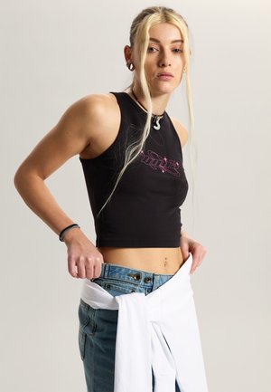 Jeune femme aux longs cheveux blonds portant un crop top noir sans manches et un jean bleu, ajustant la ceinture du jean avec les deux mains.