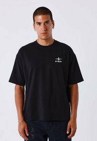Camiseta de algodón negra con mangas cortas, cuello redondo y un logo blanco en el pecho. La tela parece suave y elástica.