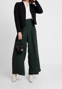 Blazer noir court porté sur une chemise blanche, associé à un pantalon vert taille haute à jambes larges avec des boutons dorés, et un petit sac à main noir texturé.