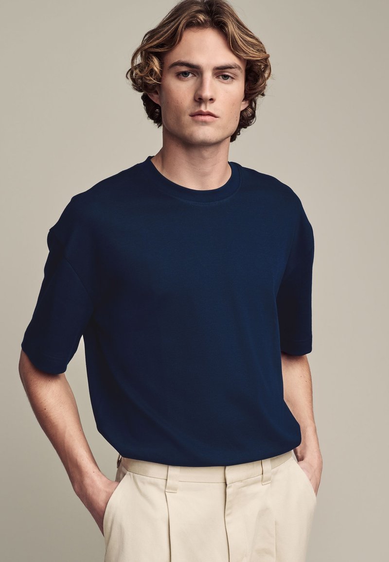 Camiseta de manga corta azul marino hecha de tela suave; presenta un cuello redondo y un corte relajado, combinada con pantalones de color claro.